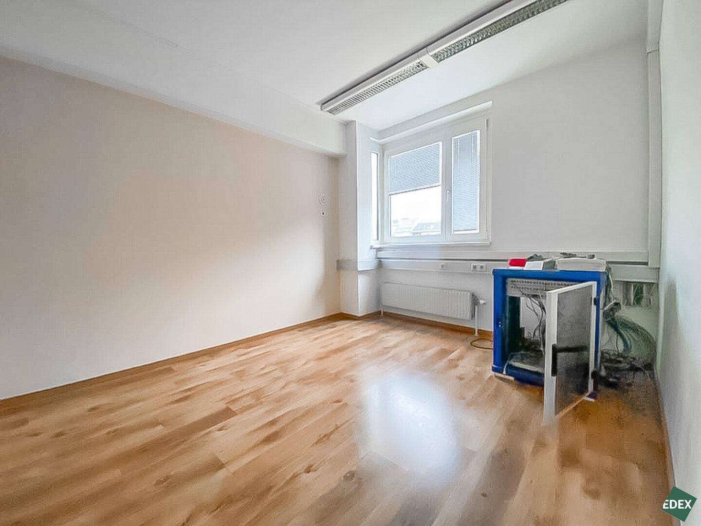 Büro mit Holzfußboden, großem Fenster und einigen technischen Geräten im Eckbereich.