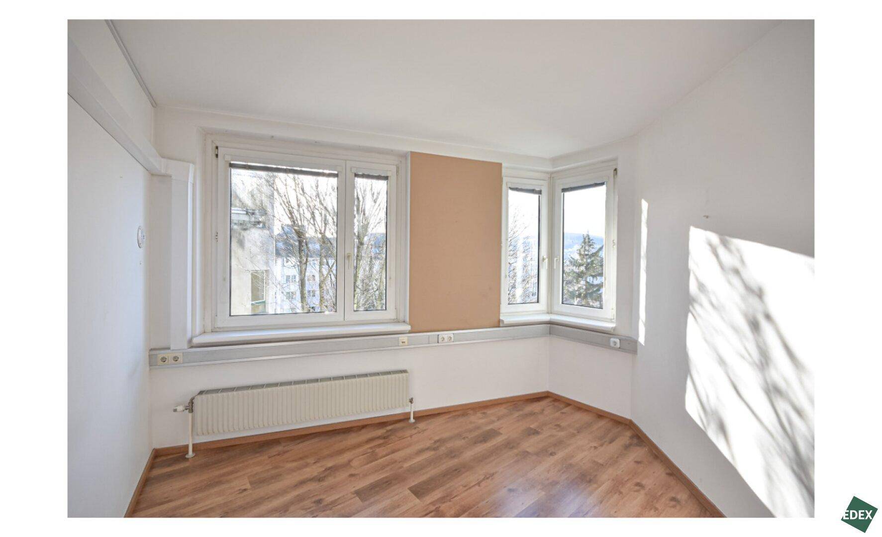 Helles Eckbüro mit großen Fenstern, Holzfußboden und einer beigen Akzentwand.