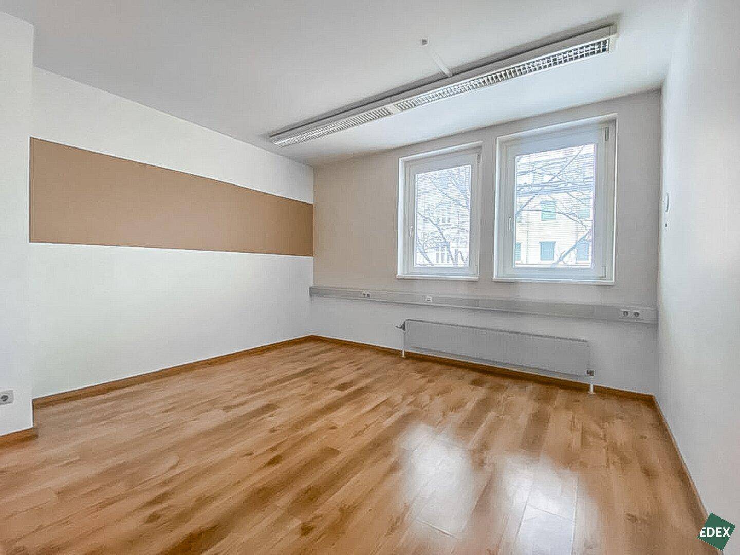 Büro mit Holzfußboden, zwei Fenstern und einer beigen Akzentwand, gut beleuchtet.