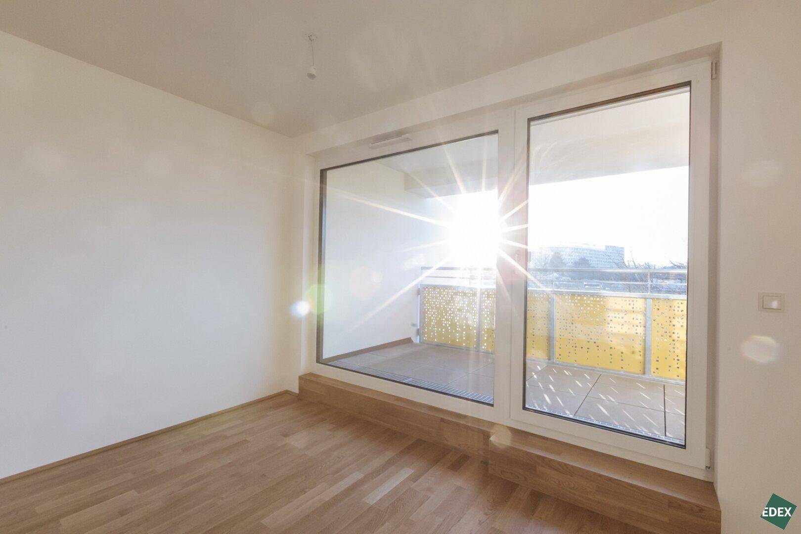 Heller Raum mit großen Fenstern und Holzboden, der viel Sonnenlicht hereinlässt.