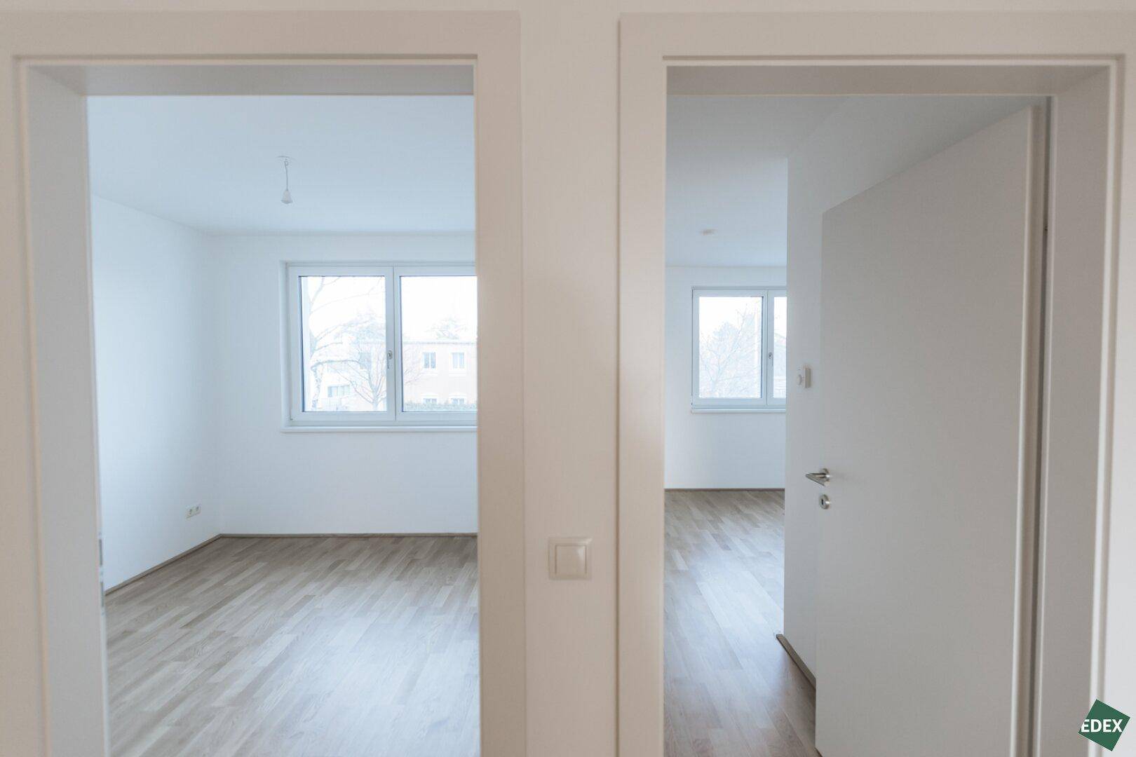 Blick vom Flur in zwei helle Zimmer mit Holzboden und Fenstern.