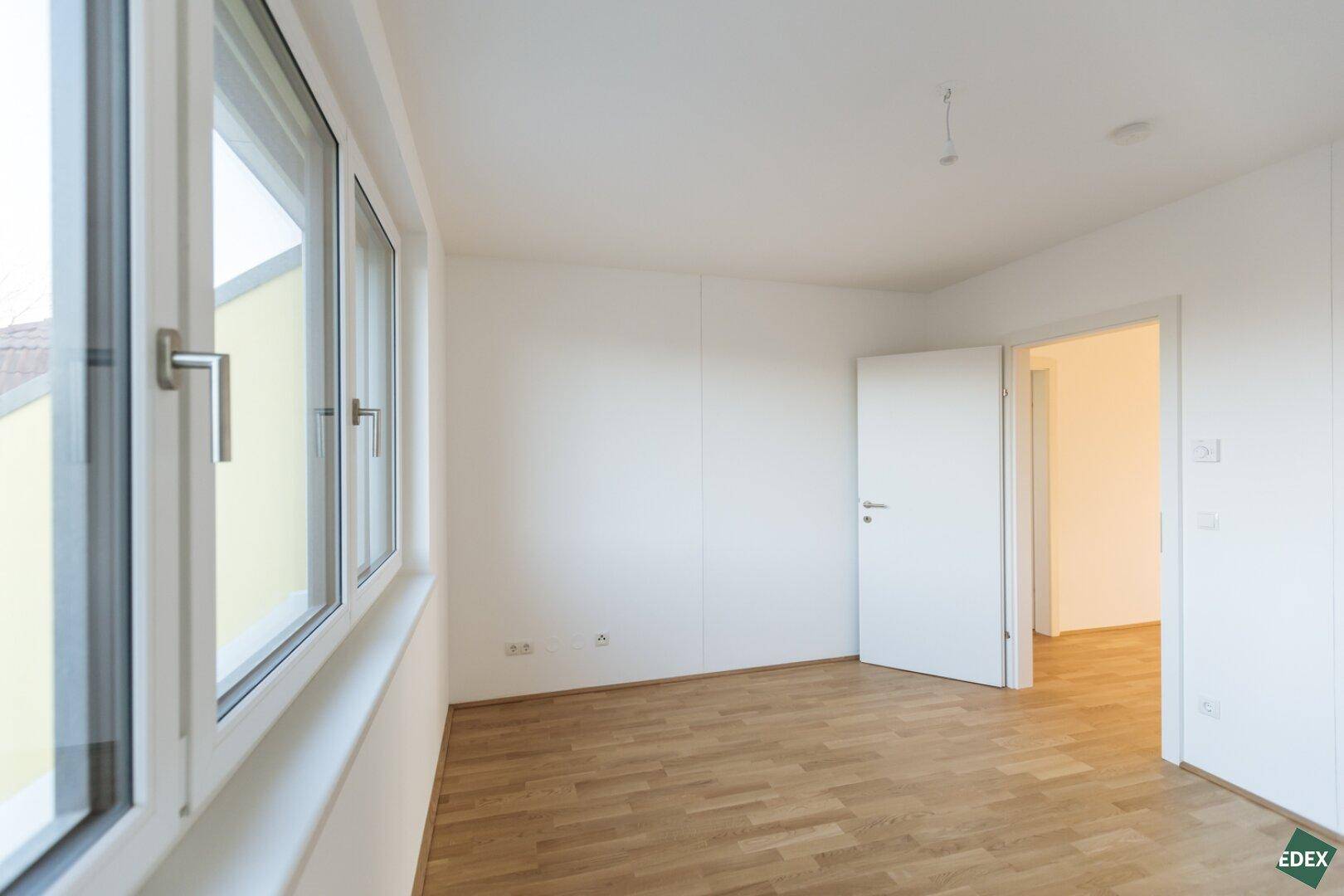 Helles Zimmer mit Holzboden, Fenster und weißer Tür.