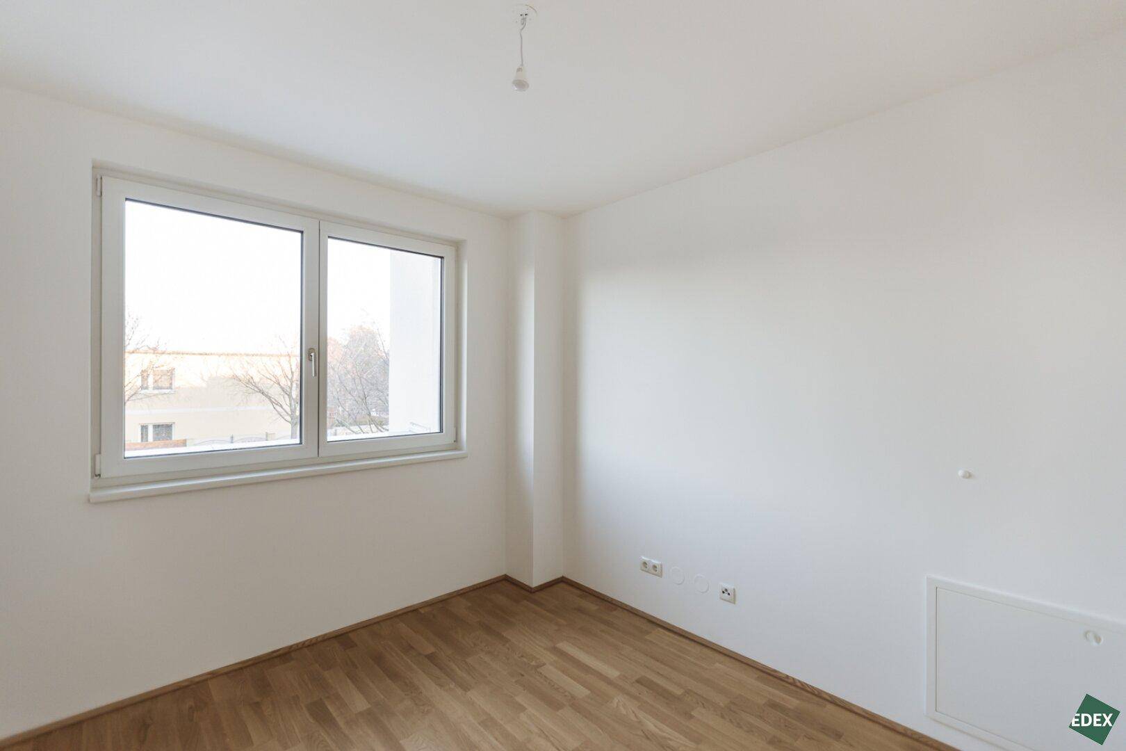 Leeres Schlafzimmer mit Holzboden und einem Fenster, das Tageslicht hereinlässt.