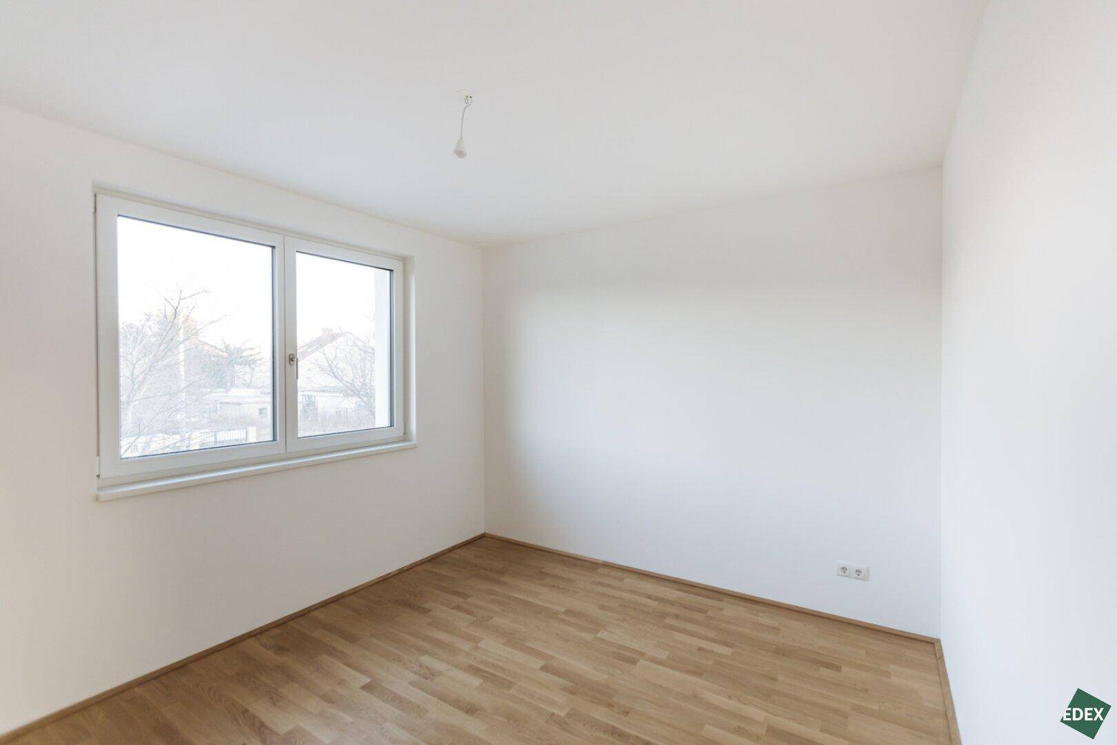 Leeres Schlafzimmer mit Holzboden und einem Fenster, das Tageslicht hereinlässt.