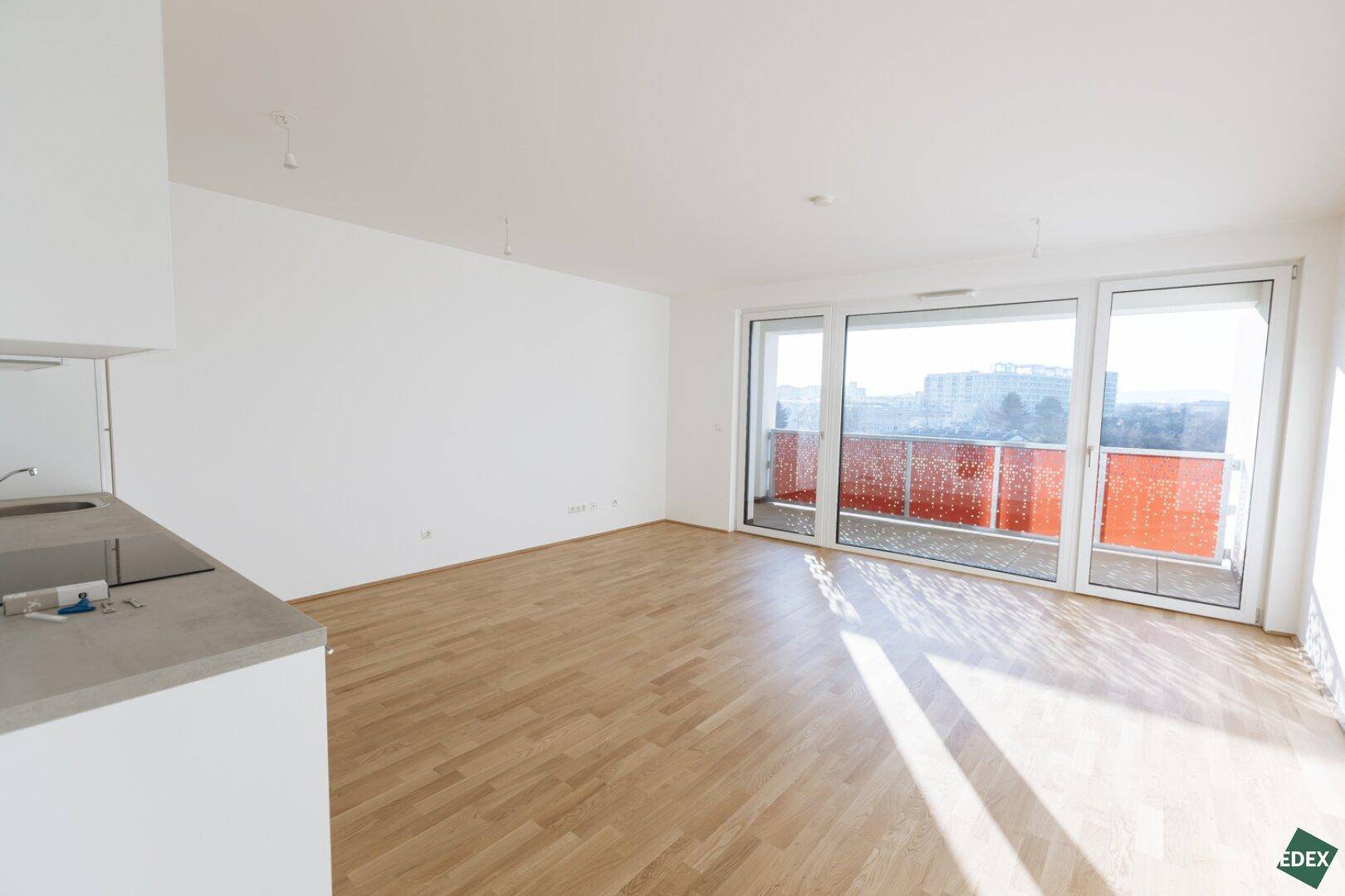 Heller, leerer Wohnbereich mit Parkettboden und großen Fenstern zum Balkon.