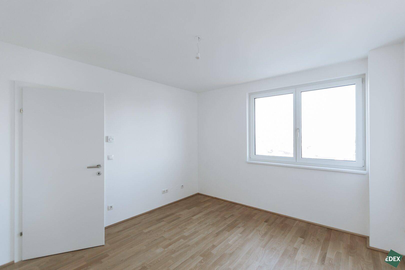 Leeres, helles Zimmer mit Parkettboden und einem großen Fenster.