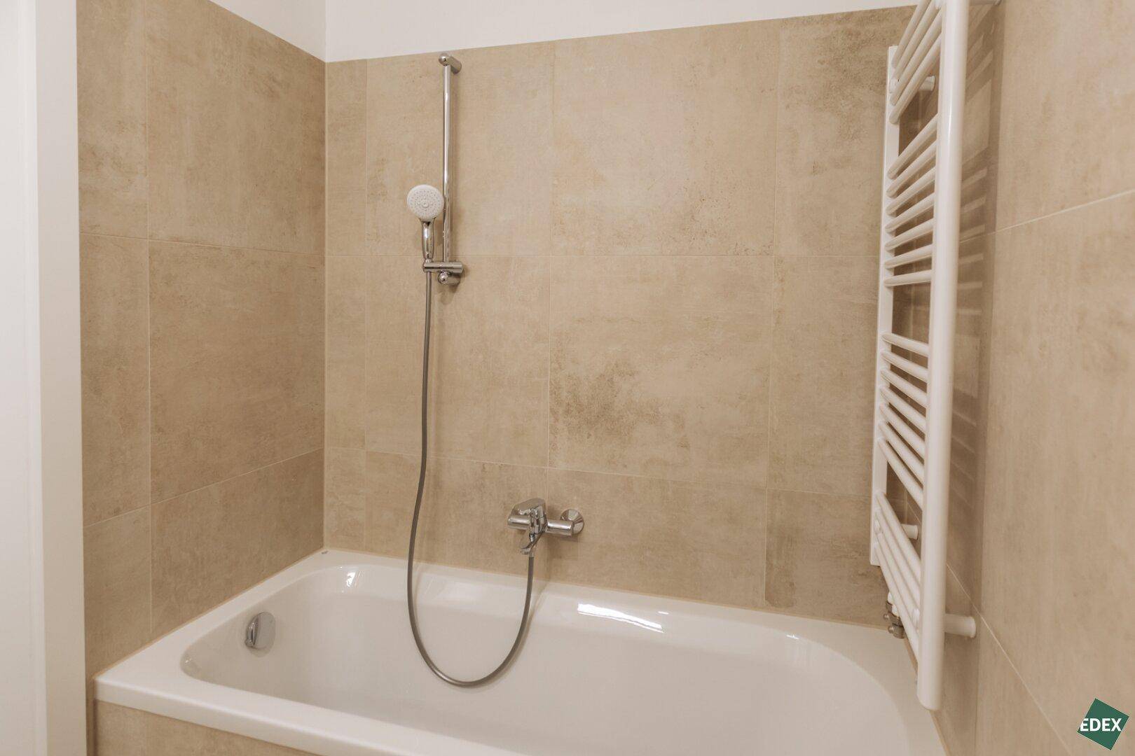 Helles Badezimmer mit Badewanne, Duscharmatur und beigefarbenen Wandfliesen.
