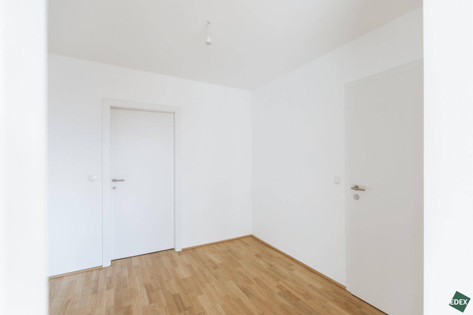 Leeres, helles Zimmer mit Parkettboden und weiß gestrichenen Wänden.