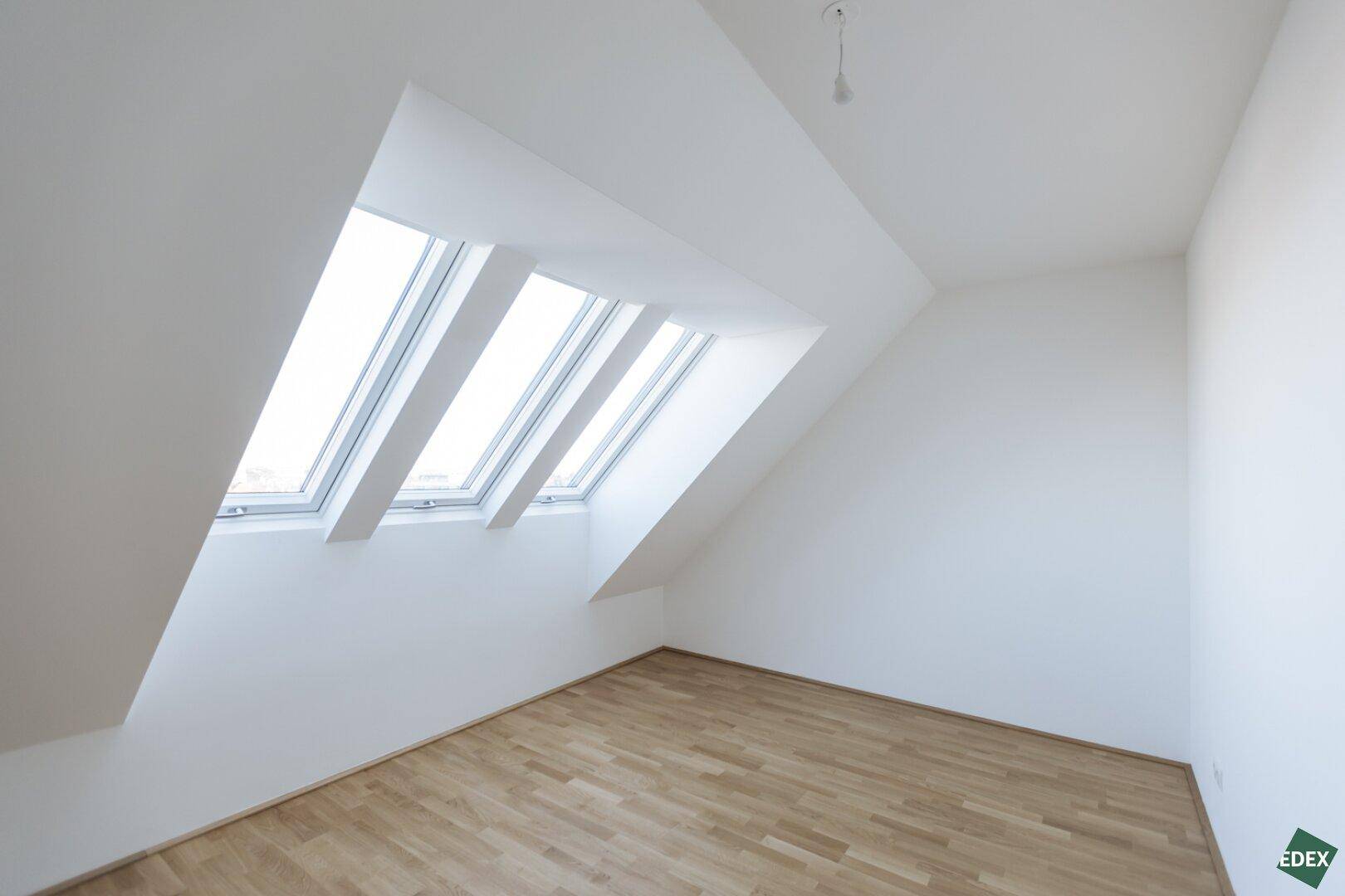 Heller Raum mit schräger Decke und drei Dachfenstern, die viel natürliches Licht hereinlassen.