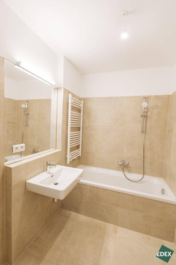 Modernes Badezimmer mit beigefarbenen Fliesen, Badewanne mit Duschfunktion, Waschbecken und Handtuchheizkörper.