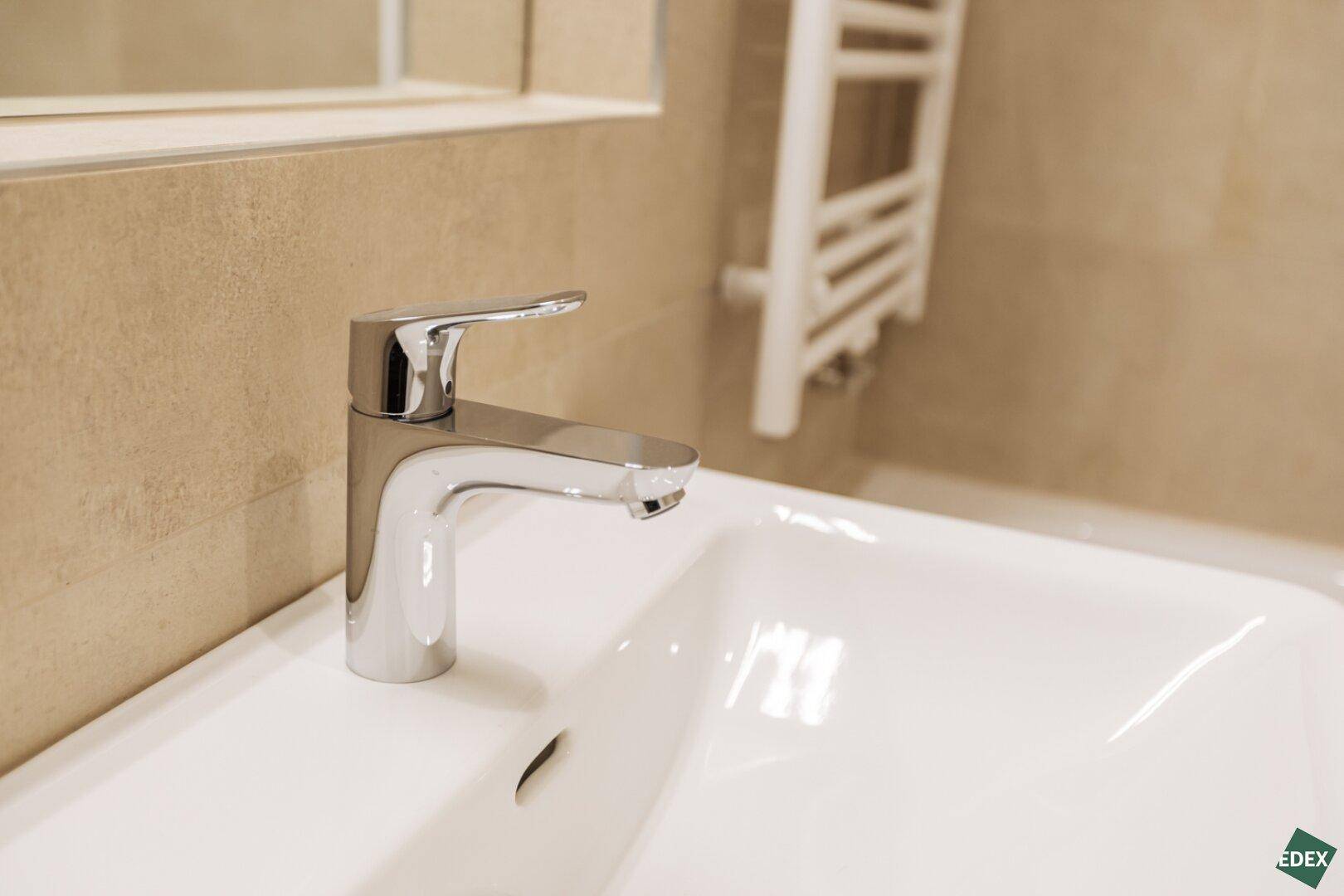 Nahaufnahme eines modernen Wasserhahns über einem weißen Waschbecken in einem gefliesten Badezimmer.