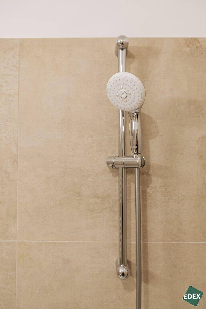 Nahaufnahme eines Duschkopfs an einer verstellbaren Stange in einem modern gefliesten Badezimmer.