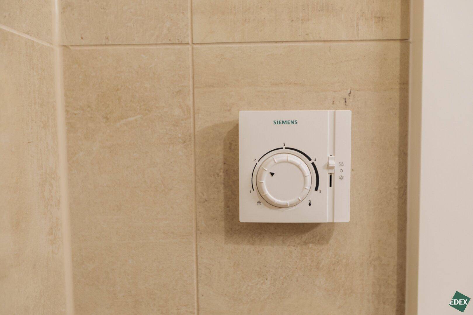 Nahaufnahme eines Siemens-Thermostats an einer beigefarbenen Fliesenwand, zur Temperaturregelung.