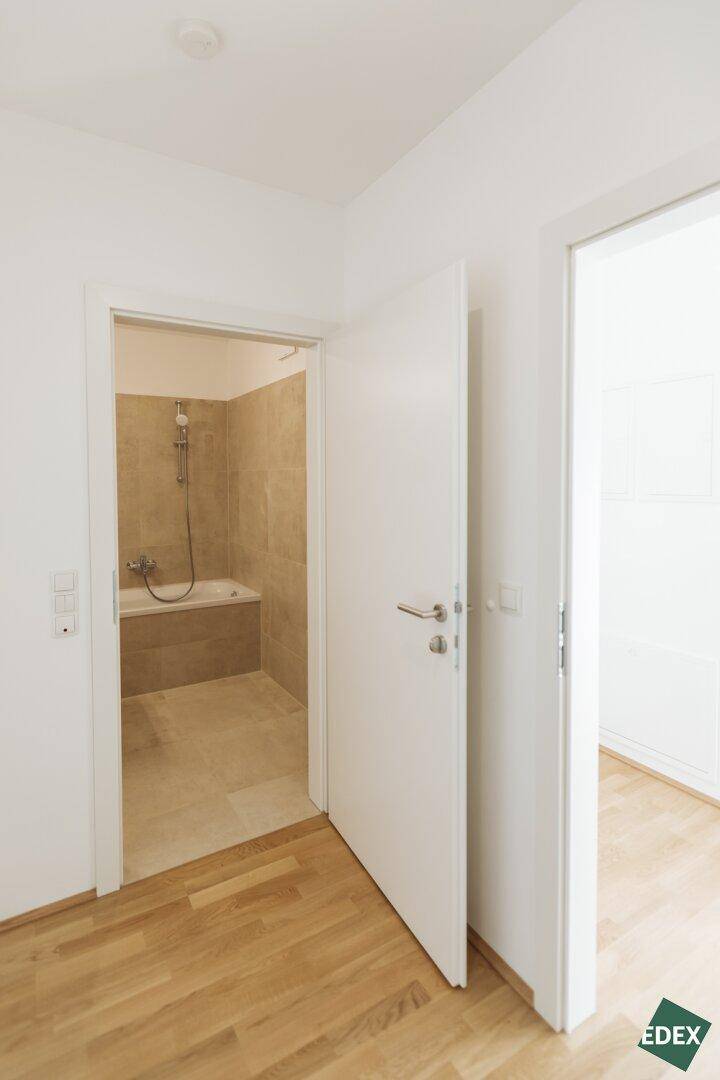 Blick vom Flur in ein Badezimmer mit Badewanne und Duschfunktion, mit Holzboden im Flur.