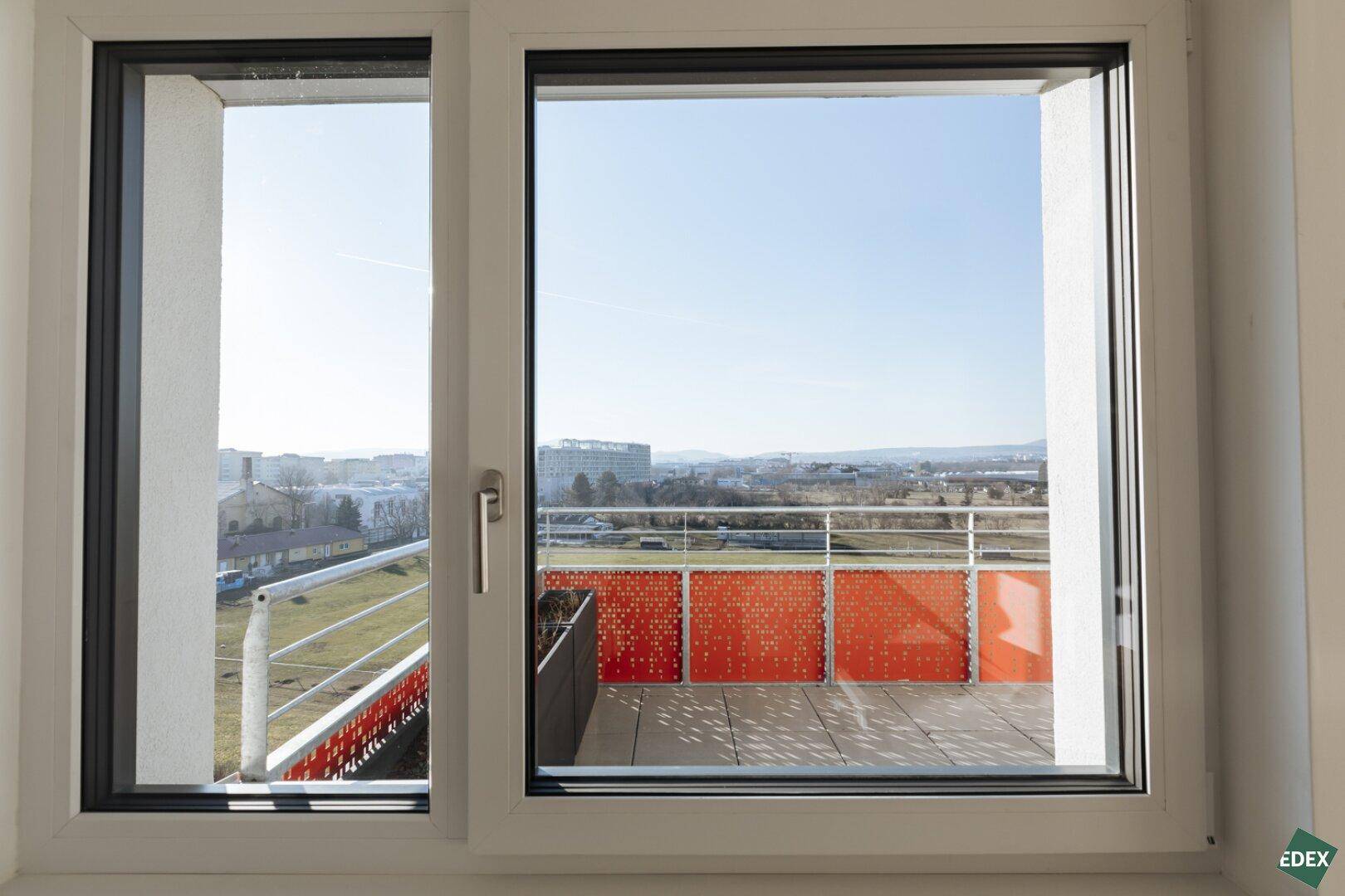Fensterblick auf eine Terrasse mit orangefarbener Brüstung und weitem Ausblick über die Stadtlandschaft.