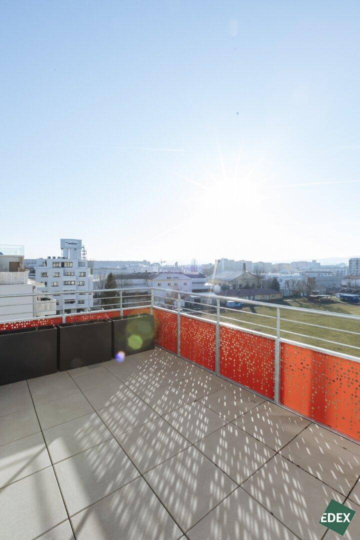 Sonnige Terrasse mit moderner orangefarbener Brüstung und weitem Blick über die städtische Umgebung.