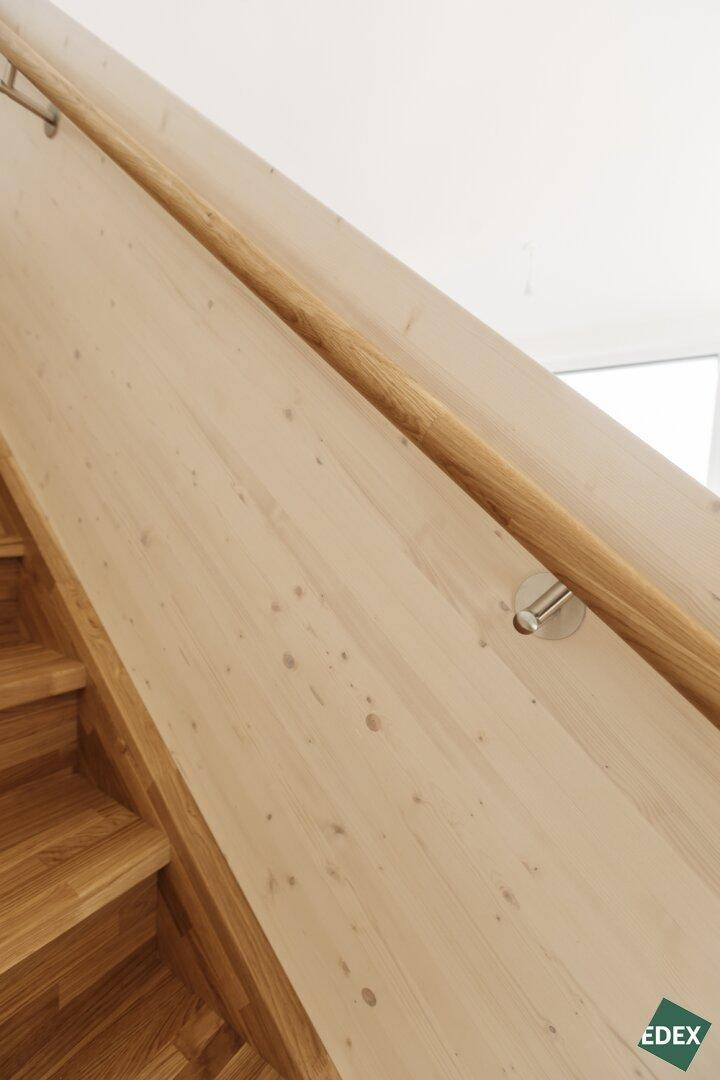 Nahaufnahme eines hölzernen Handlaufs an einer hellen Holzwand neben einer Treppe.