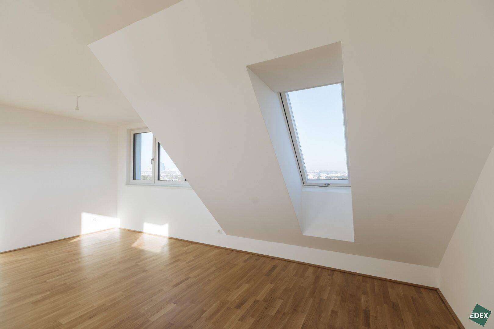 Lichtdurchfluteter Dachgeschossraum mit schrägen Wänden, Dachfenster und Blick auf die Stadt.