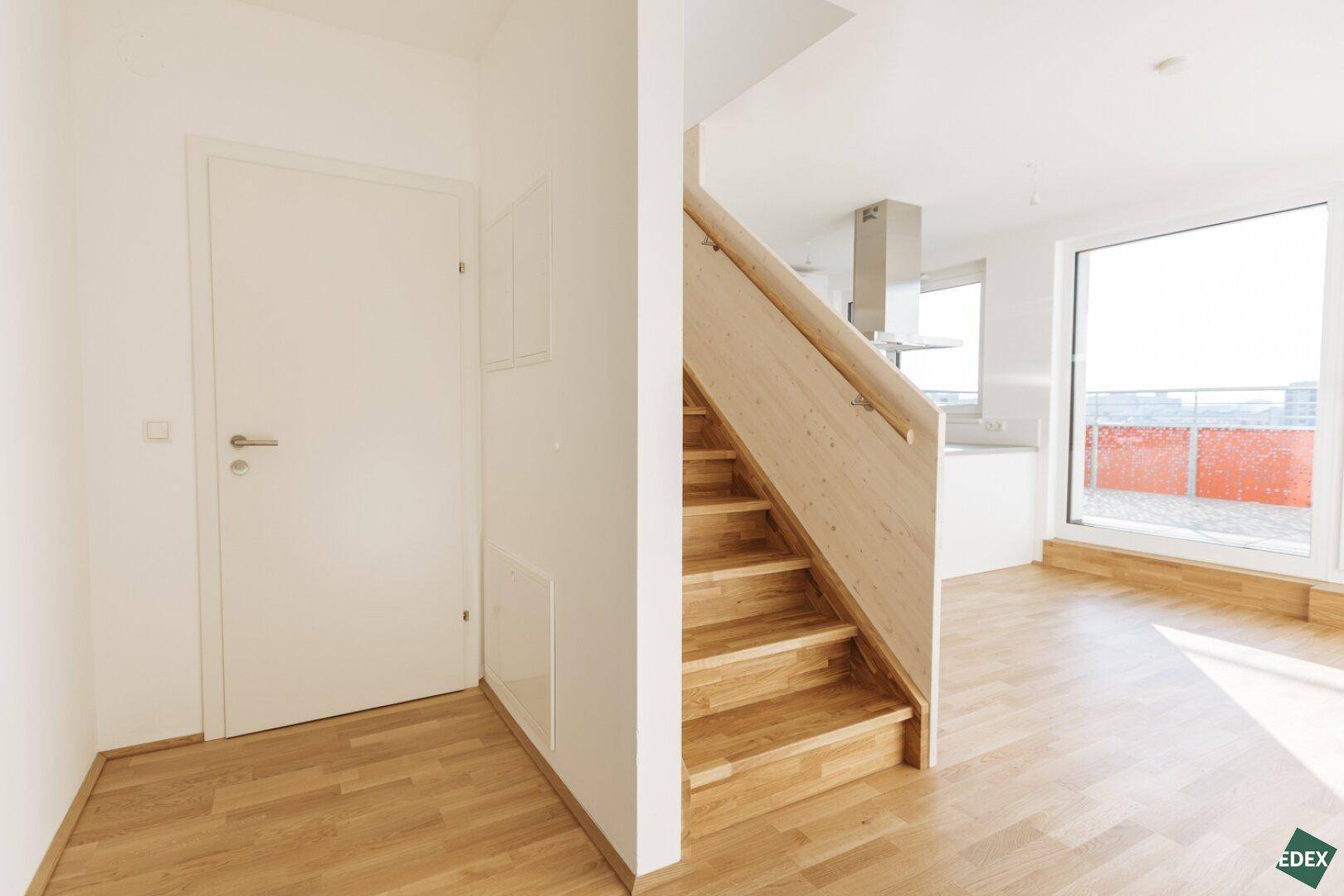 Heller Wohnbereich mit Holztreppe und Zugang zu einem Balkon, offene Küche im Hintergrund sichtbar.