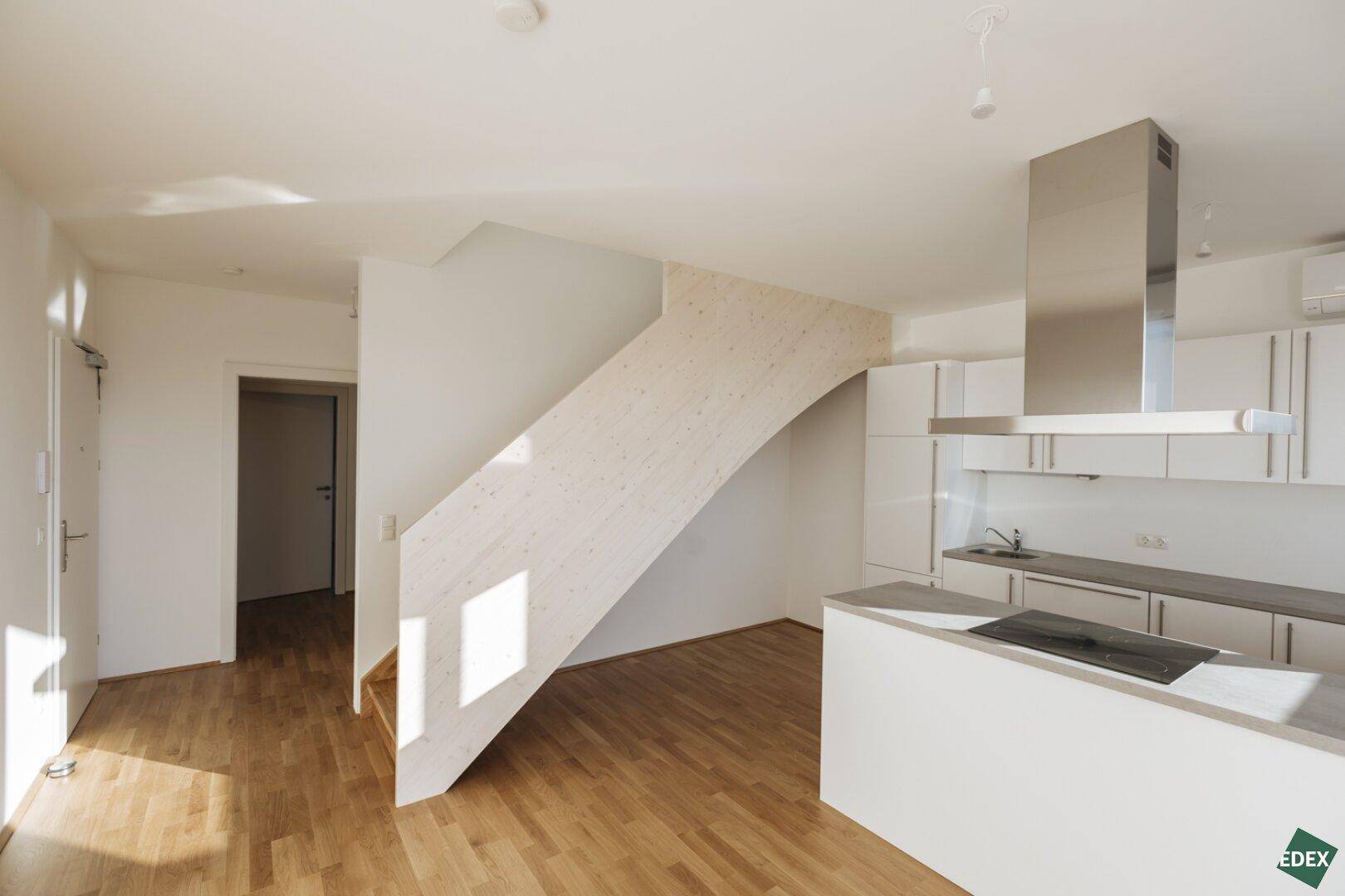 Heller offener Wohnbereich mit moderner Einbauküche, Holztreppe und Parkettboden.