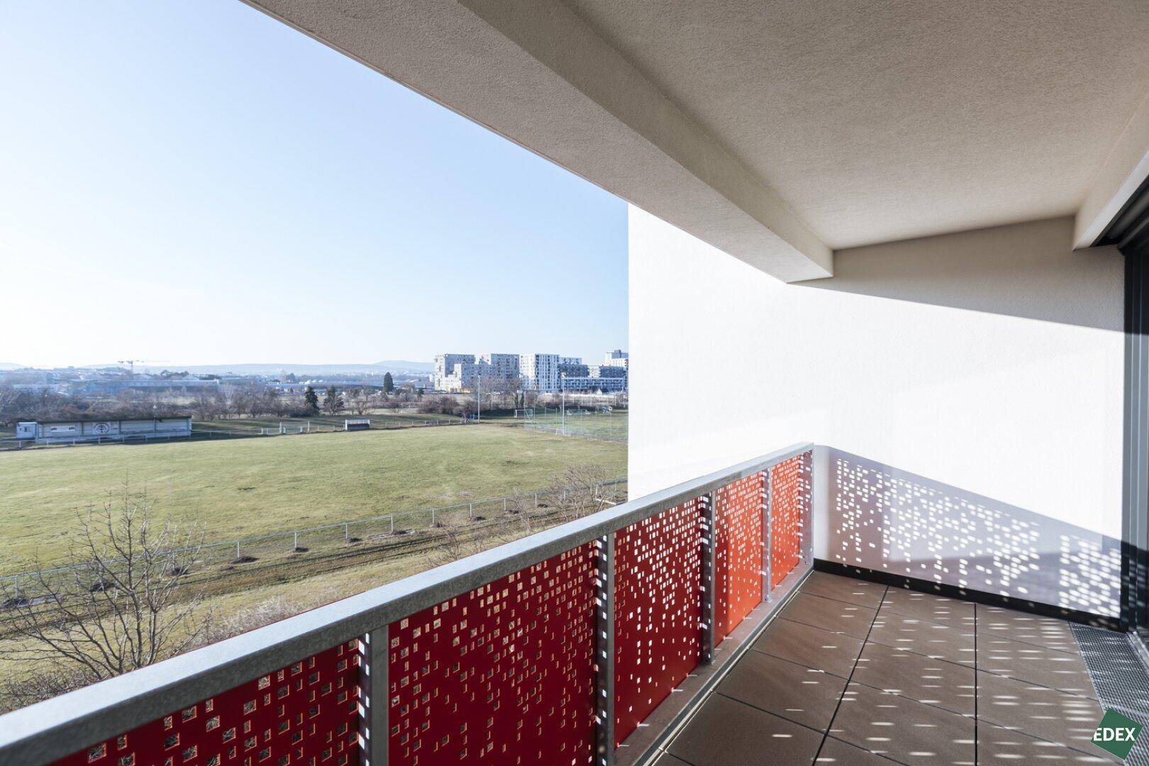 Schmaler Balkon mit rotem Lochblechgeländer und Blick auf grüne Wiesen und die Stadt.