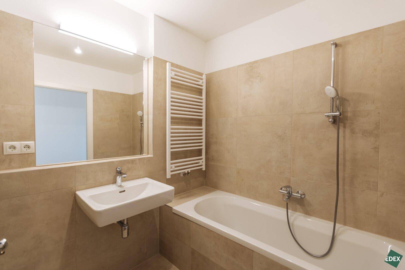 Modernes Badezimmer mit beigefarbenen Fliesen, Badewanne, Waschbecken und Handtuchheizkörper.