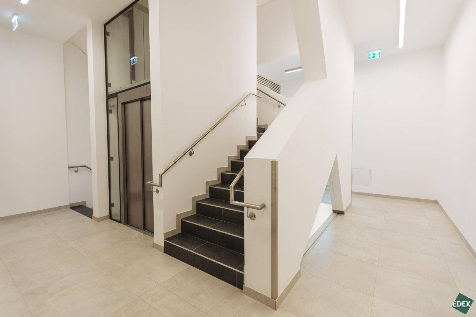 Moderner Eingangsbereich mit Treppe, Aufzug und hellen Fliesen im Mehrfamilienhaus.
