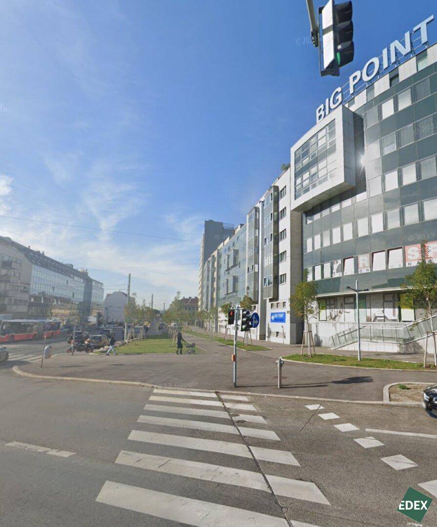 Modernes Geschäftsgebäude an einer belebten Straße mit Fußgängerüberweg und klarem Himmel, umgeben von städtischer Architektur.