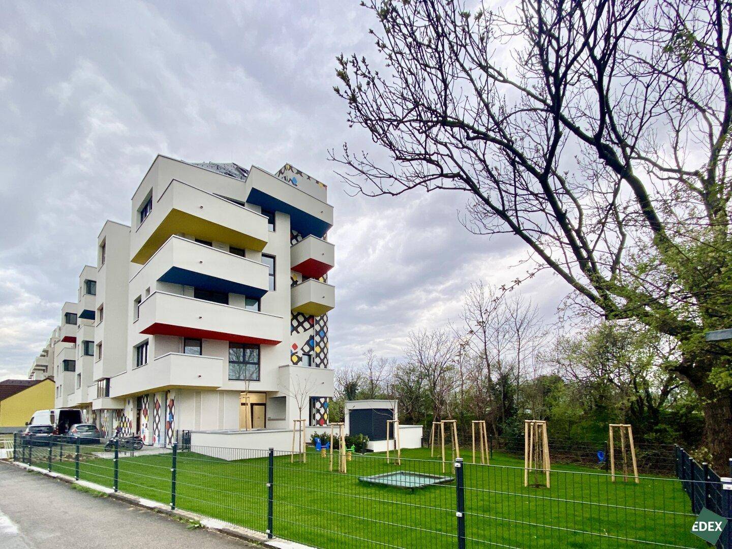 Modernes Mehrfamilienhaus mit farbigen Balkonen und einer Grünfläche mit Spielbereich unter bewölktem Himmel.