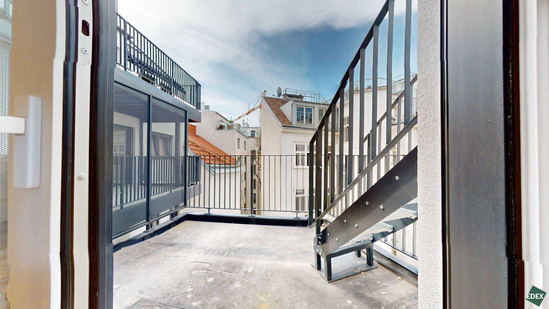 Geräumige Dachterrasse mit Metallgeländer und Treppe, umgeben von städtischen Gebäuden.