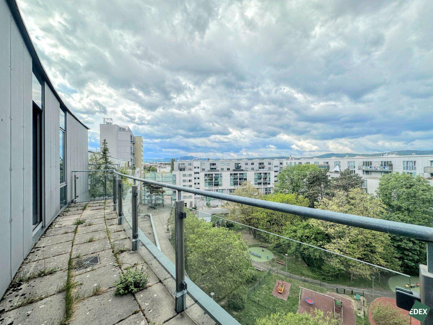 Langer, geschwungener Balkon mit Glasgeländer und Blick auf eine Wohnsiedlung mit Grünflächen und Spielplatz.