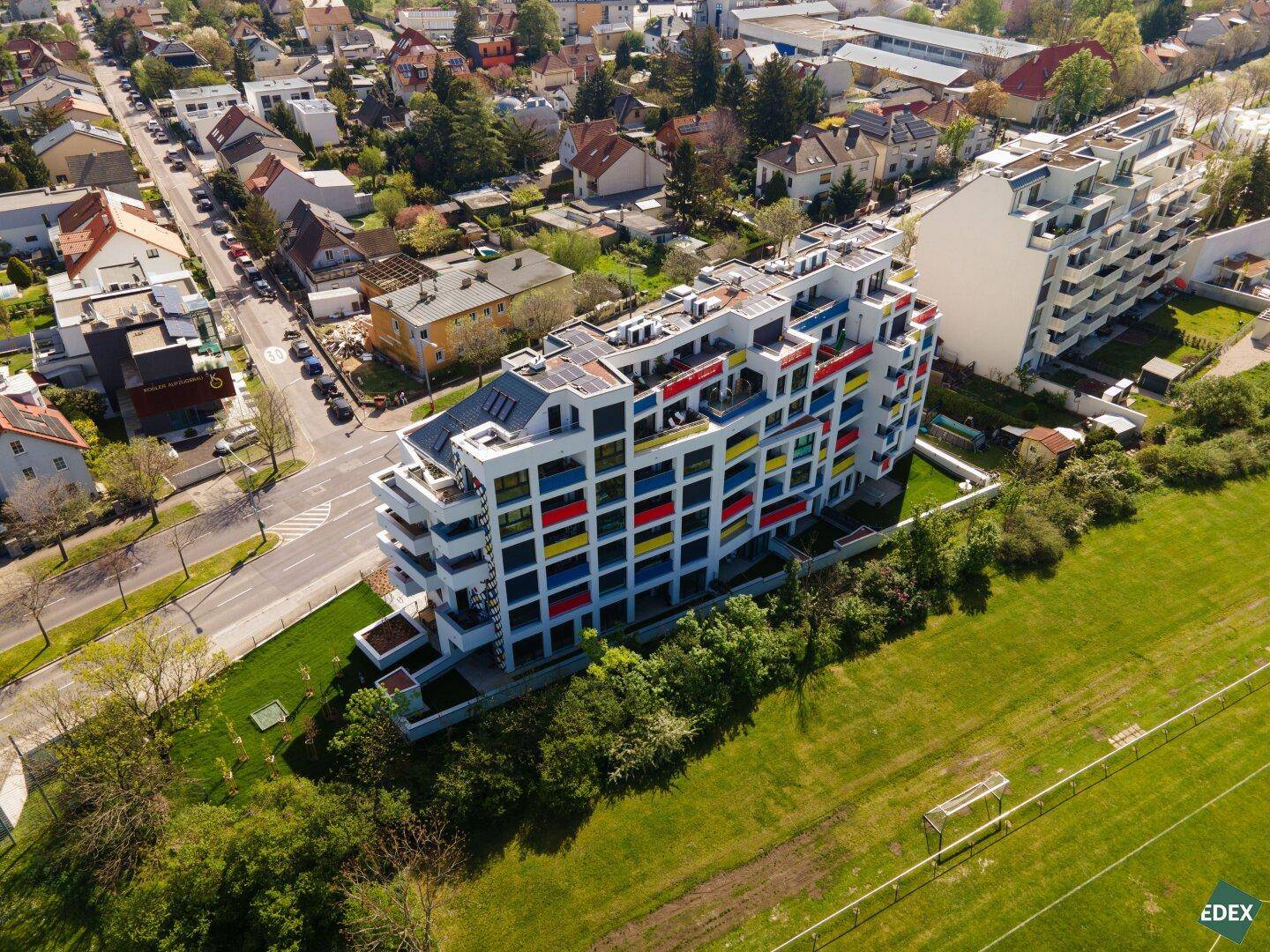 Schräge Luftaufnahme des modernen Wohngebäudes mit farbigen Balkonen und angrenzendem Sportplatz.