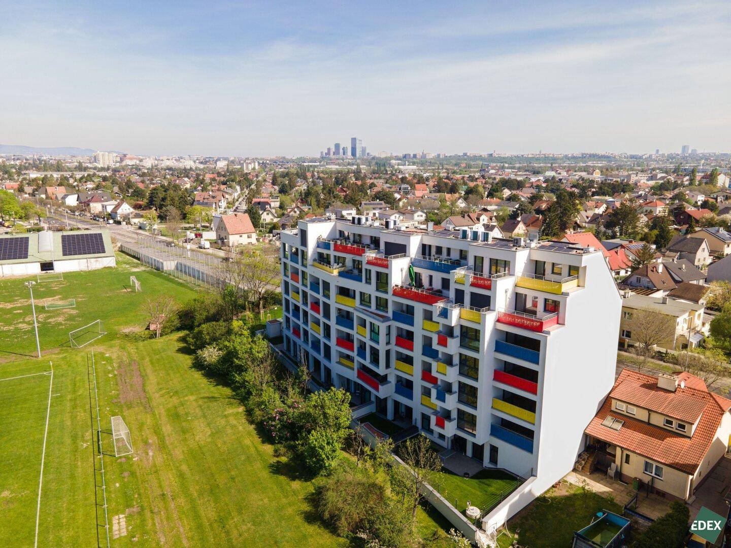 Weitläufige Luftaufnahme der Wohnanlage mit farbigen Balkonen, angrenzenden Sportfeldern und Stadtpanorama.