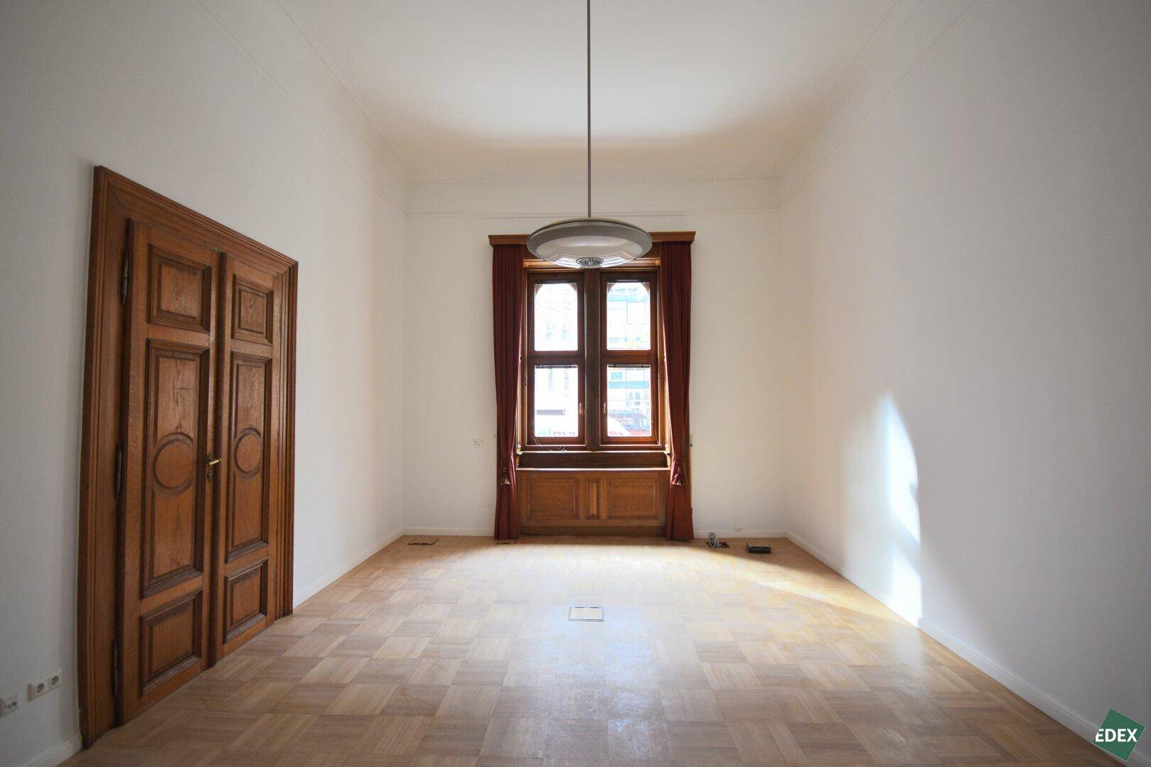 Leerer Raum mit hellem Parkettboden, einer geschlossenen Holztür und einem großen Fenster.