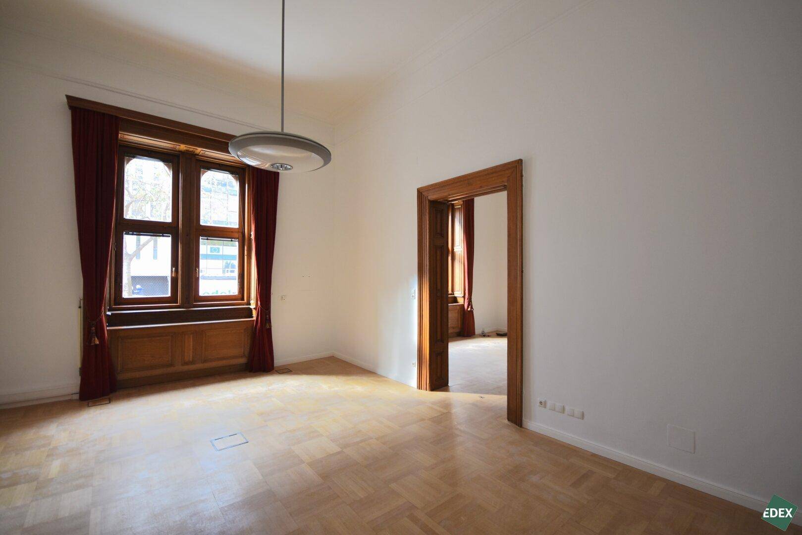 Heller Raum mit Parkettboden, einem großen Fenster und einer offenen Holztür zu einem Nebenraum.