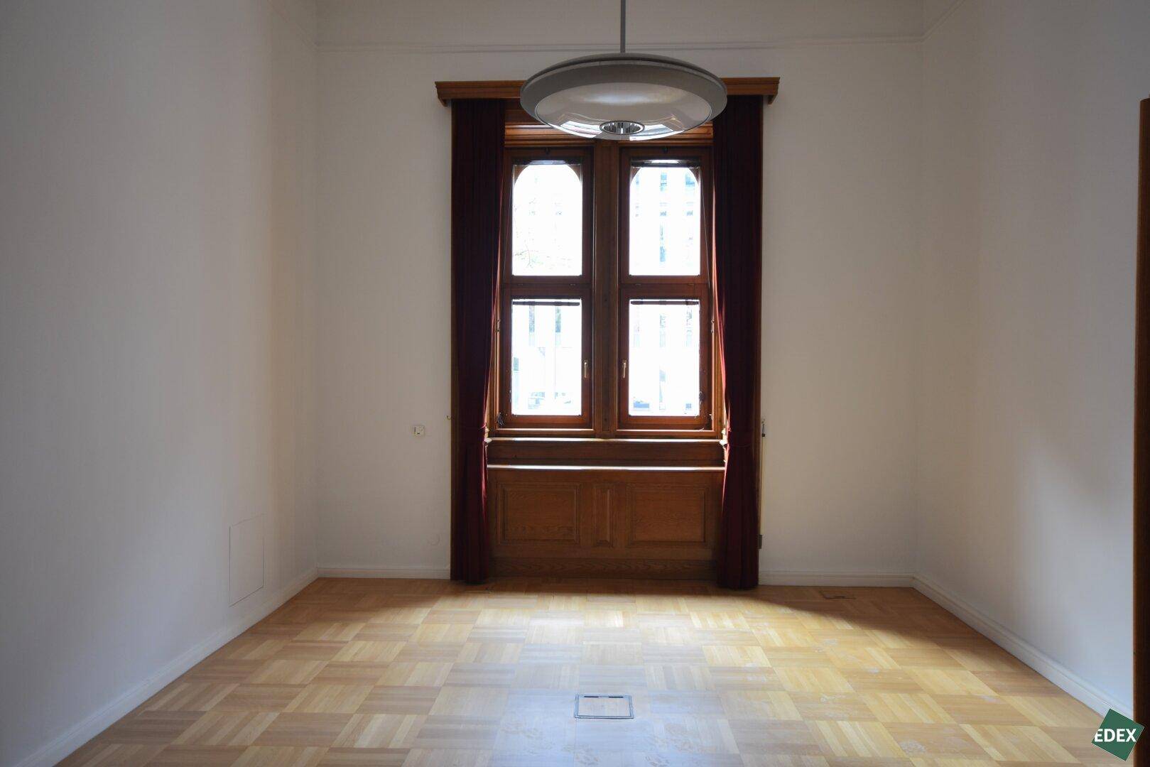 Leerer Raum mit Parkettboden und einem großen, mehrteiligen Holzfenster, das viel Licht hereinlässt.