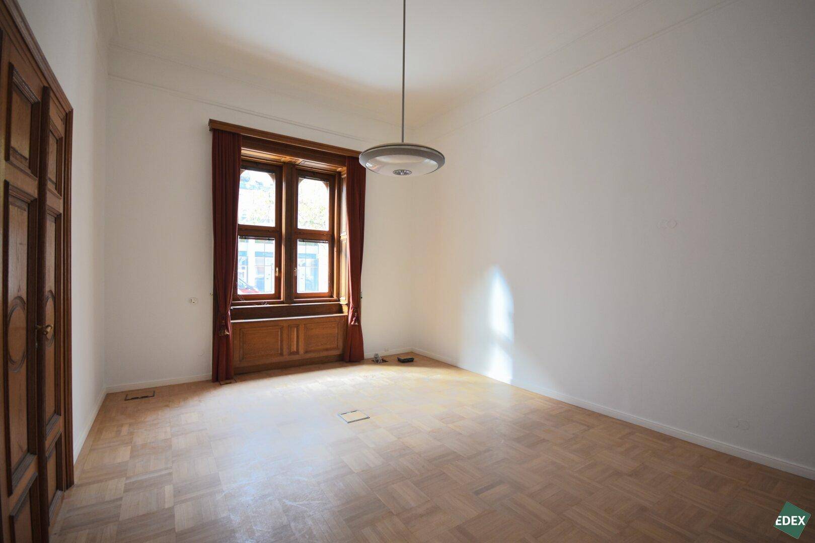 Geräumiger leerer Raum mit Parkettboden, einem großen Fenster und einer geschlossenen Holztür.