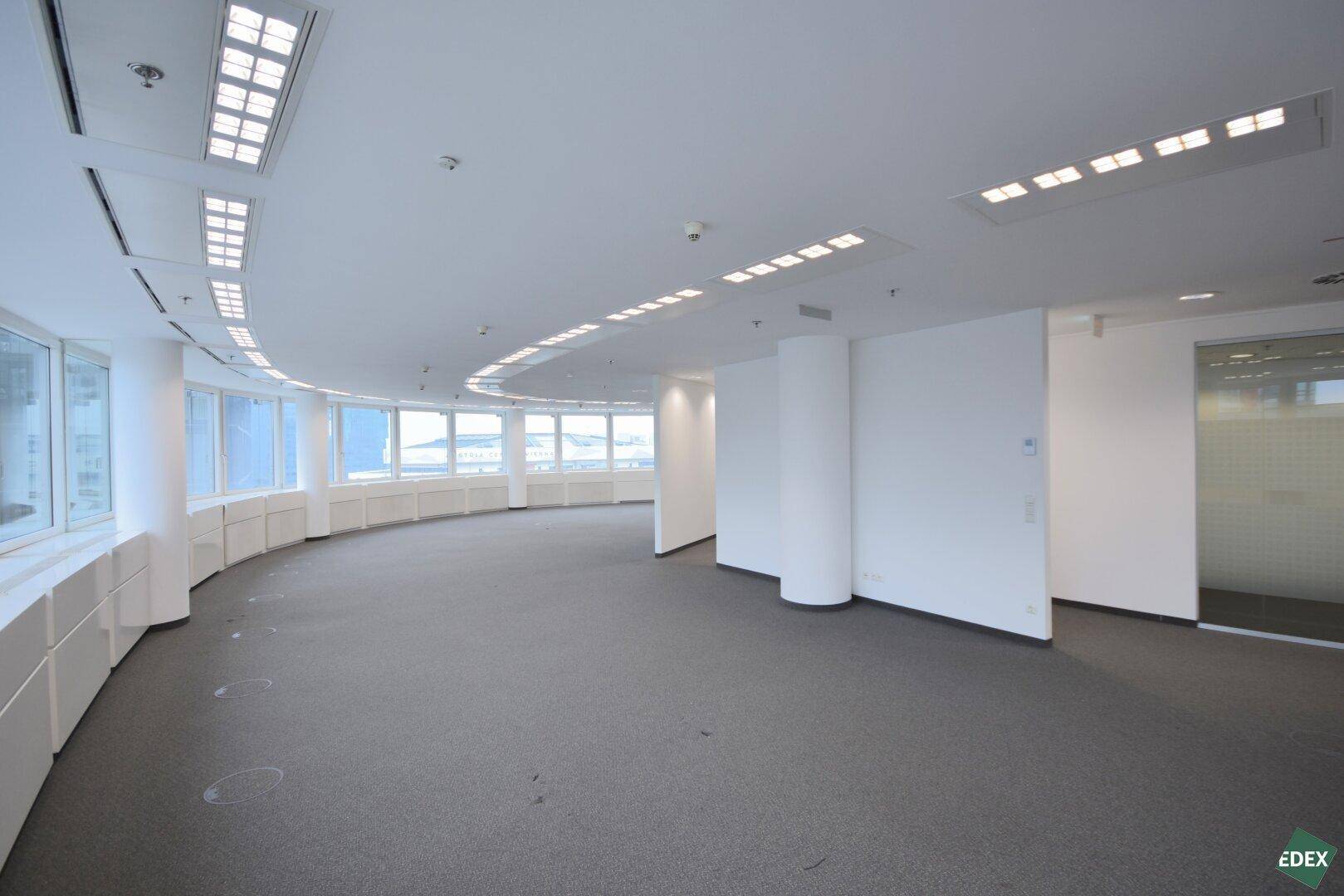 Weitläufiger Bürobereich mit Teppichboden, vielen Fenstern und moderner Deckenbeleuchtung in einem Hochhaus.
