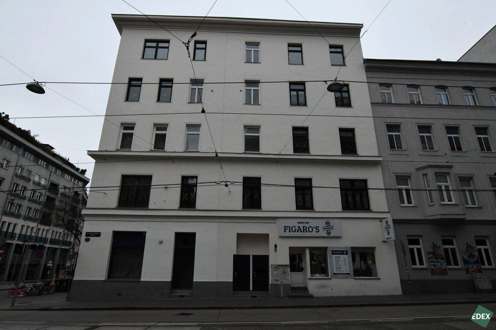 Mehrstöckiges Gebäude mit weißer Fassade und Geschäftsräumen im Erdgeschoss an einer Straße.