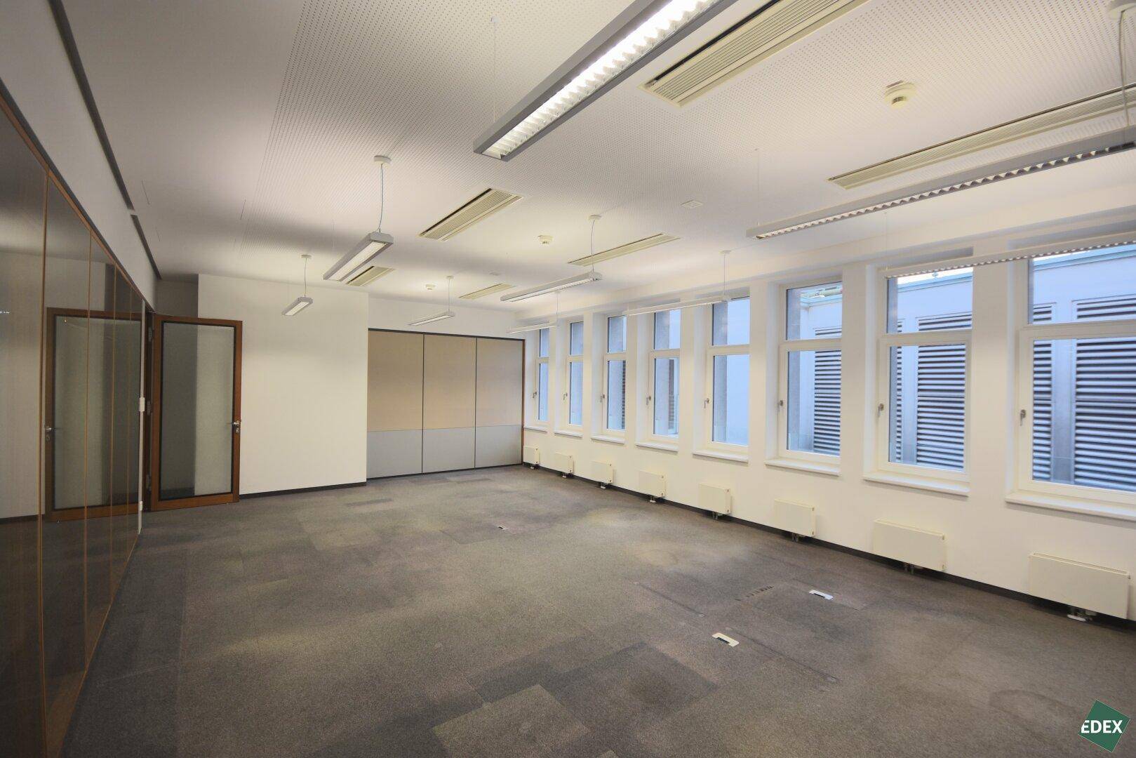 Geräumiges Büro mit Teppichboden und großen Fenstern, ausgestattet mit modernen Deckenleuchten und Klimaanlage.
