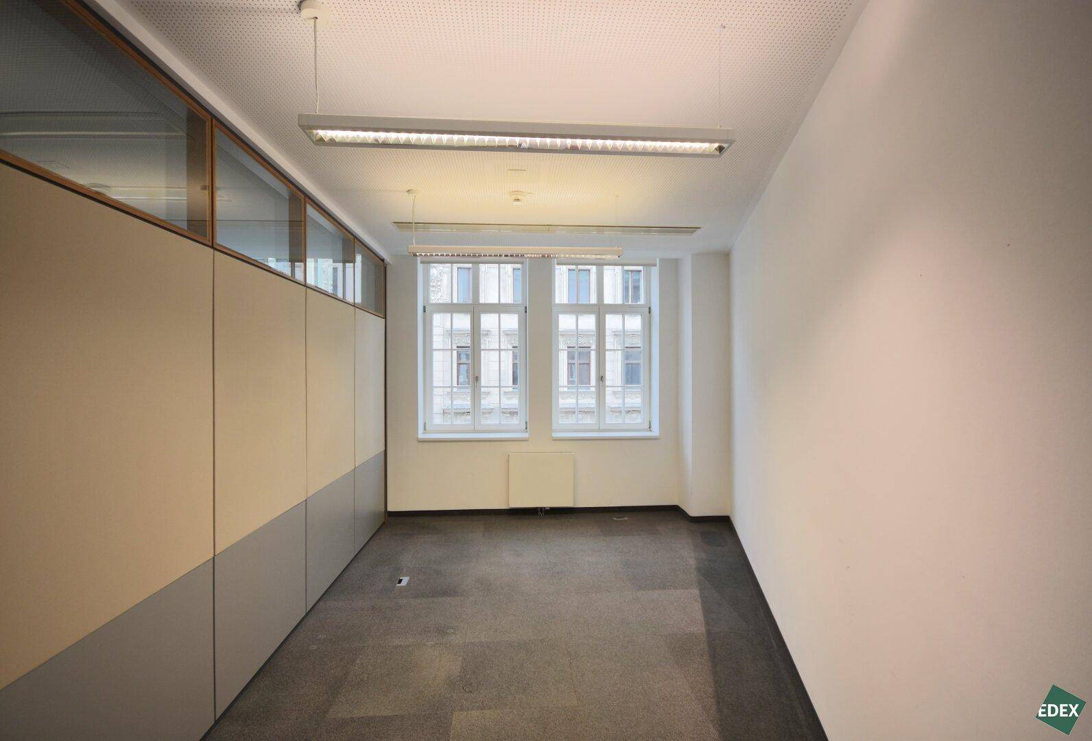 Kleines Büro mit zwei Fenstern und Teppichboden, bietet eine ruhige Arbeitsumgebung mit Tageslicht.