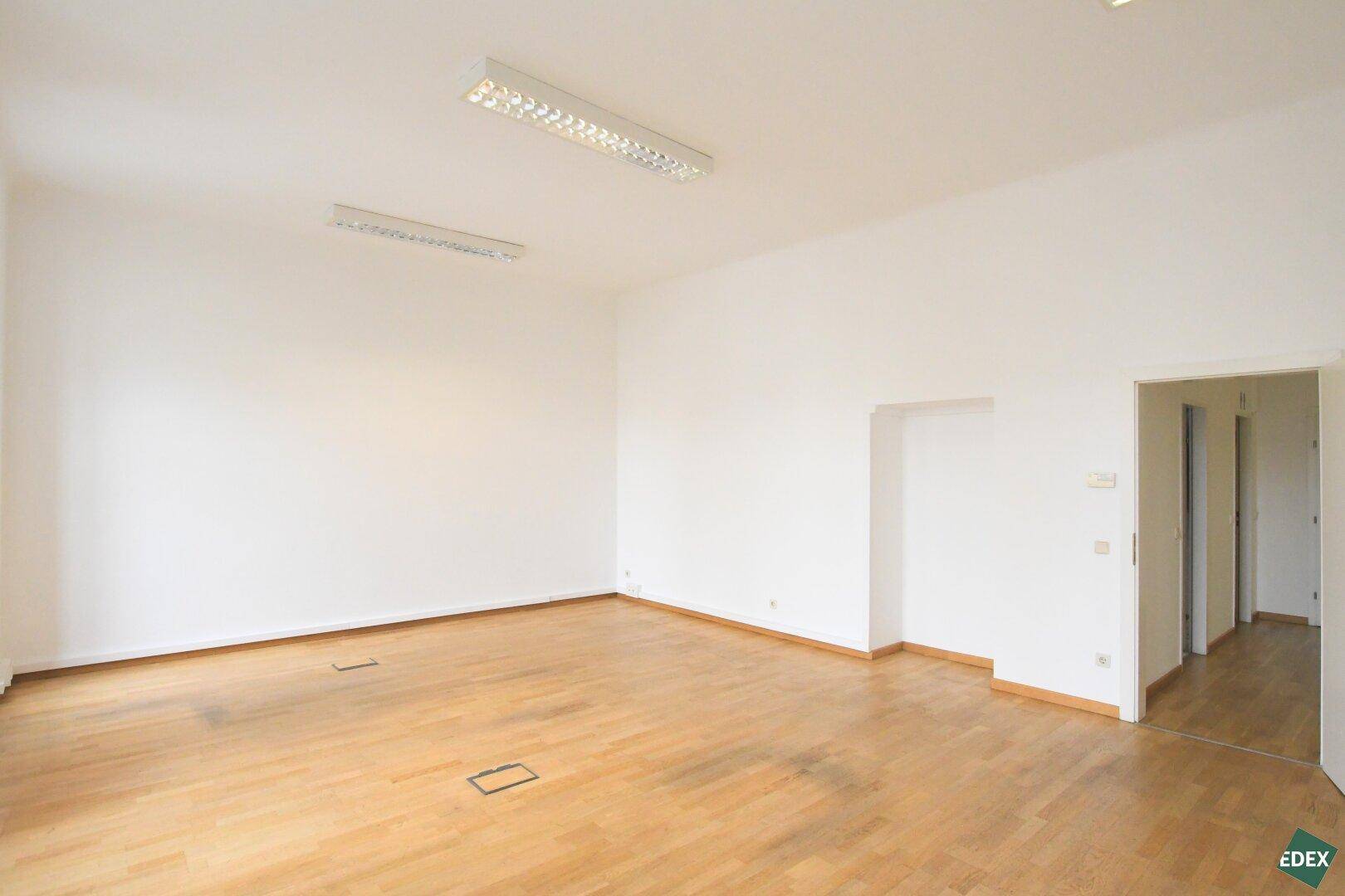 Geräumiger, heller Büroraum mit Holzboden und Zugang zu weiteren Räumen durch eine Tür.