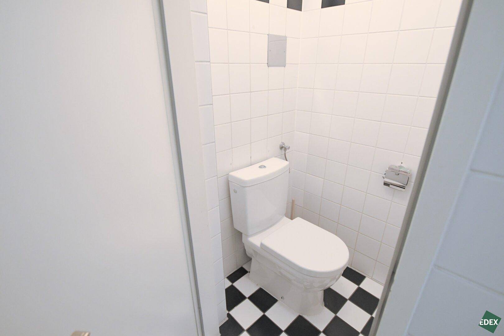 Kleine Toilette mit weißer Keramik, schwarz-weiß kariertem Fliesenboden und weißen Wandfliesen.