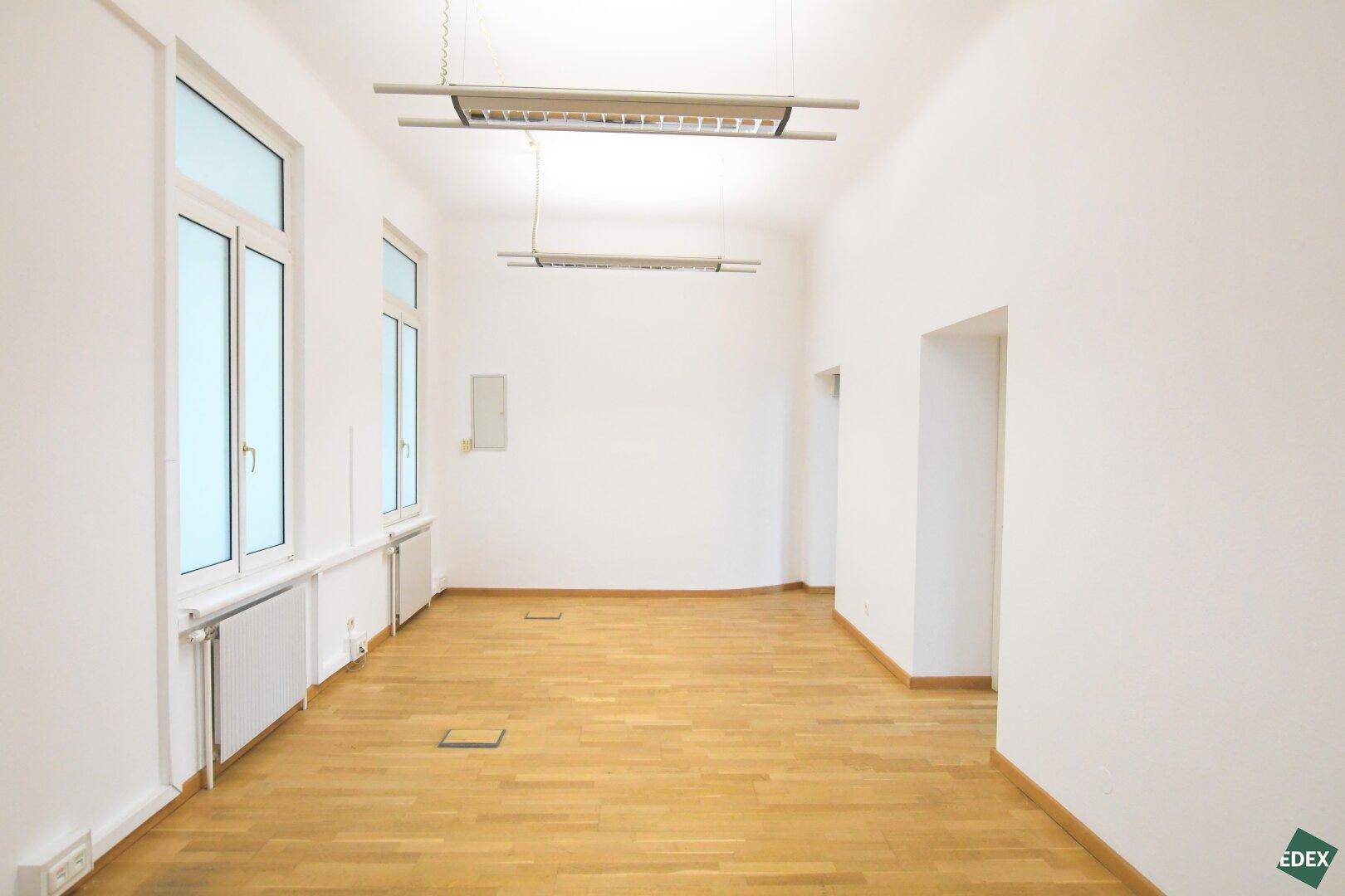 Langer, heller Raum mit Holzboden, hohen Decken und zwei großen Fenstern, ideal als Büro.