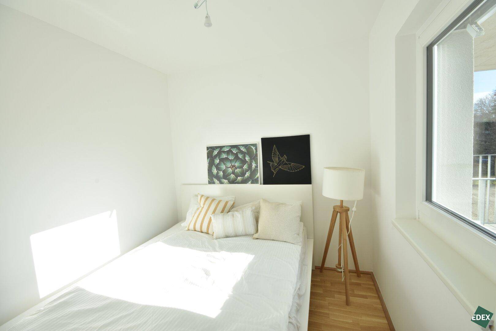 Helles Schlafzimmer mit Holzboden, großem Fenster und minimalistischer Einrichtung, sonnig und ruhig.