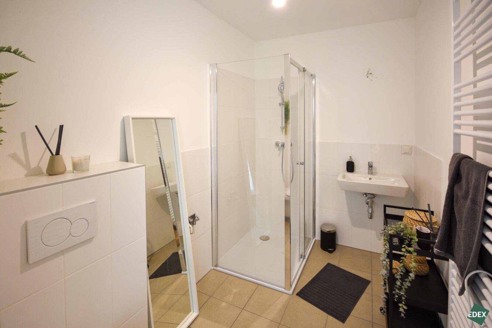 Modernes Badezimmer mit Dusche, Waschbecken und beheiztem Handtuchhalter, hell gefliest.