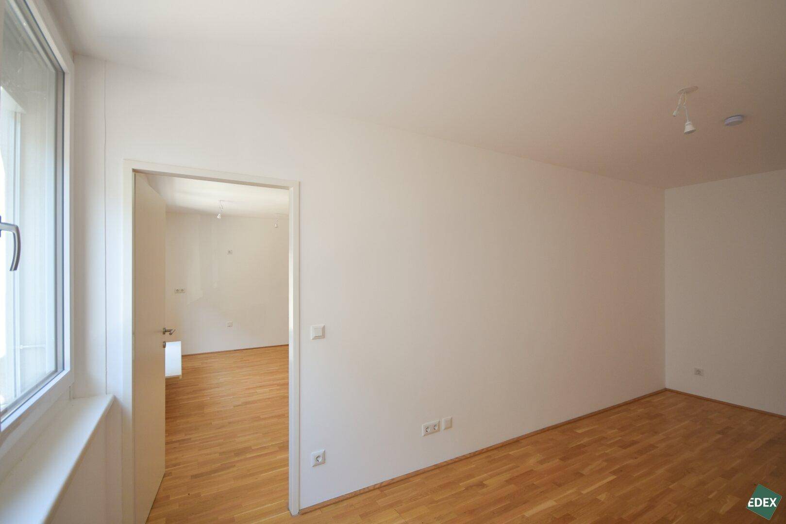 Heller Raum mit Parkettboden und offener Tür zu einem weiteren Zimmer, frisch gestrichen.