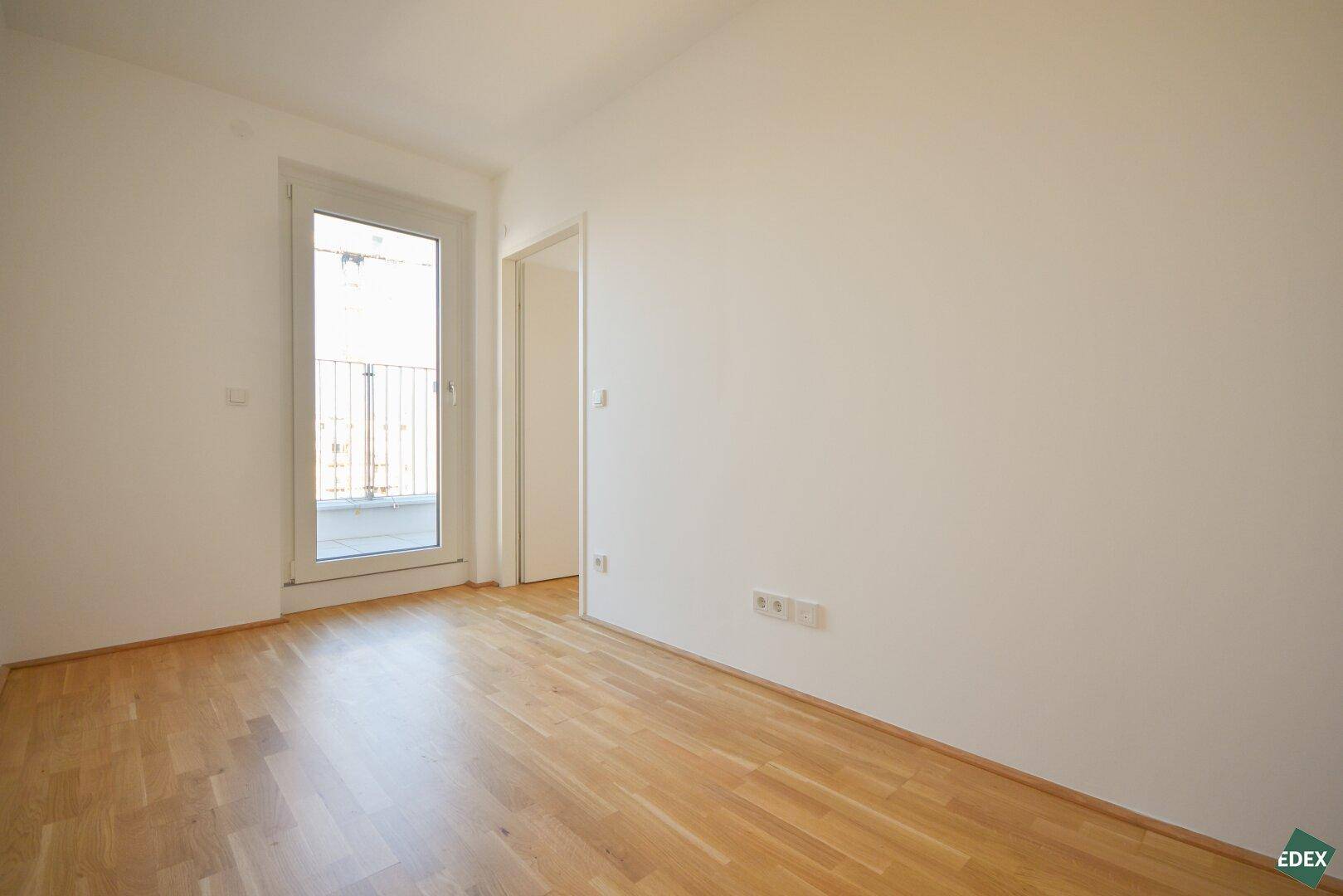 Leeres Schlafzimmer mit Holzboden und Zugang zu einem kleinen Balkon.