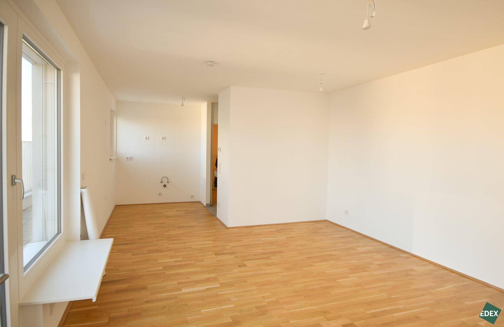Heller, leerer Wohnbereich mit Holzboden und vorbereiteten Anschlüssen für eine Küche.