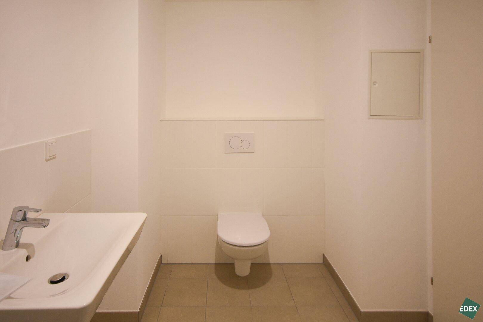 Separates WC mit wandhängender Toilette, Waschbecken und hellen Fliesen.
